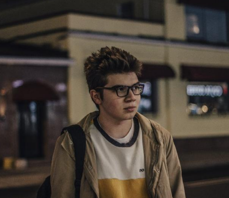 Person wearing glasses and a beige jacket standing in front of a train at night.