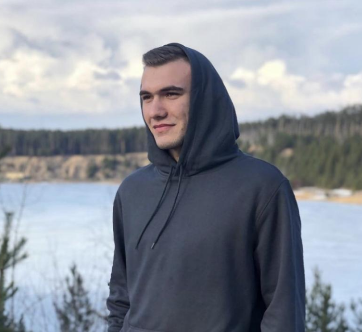 Person wearing a dark hoodie standing by a lake with trees in the background